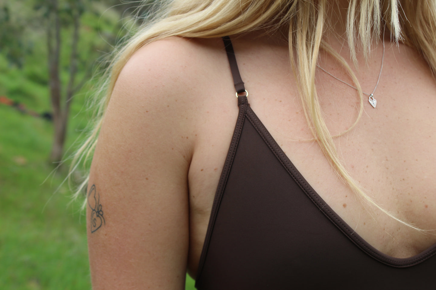 Close-up of a person wearing a brown one piece swimsuit with gold hardware with a blurred outdoor background
